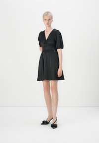 Vestido preto com decote em V, mangas curtas bufantes e cintura elástica. Tecido suave com saia rodada. Usado com sapatos pretos de salto alto com detalhe de laço.