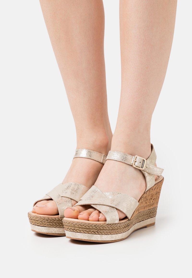 Sandales compensées beiges avec une base en liège texturée, des brides métalliques, une boucle à la cheville et un design à bouts ouverts.