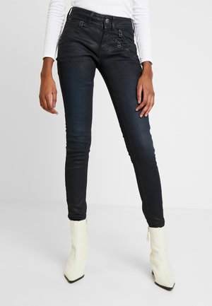 Jeans negros ajustados con bolsillos con cremalleras y una textura suave, combinados con botas de tacón alto blancas, de punta y tobilleras.
