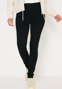 Jeans ajustados negros de denim elástico, con cintura alta, bolsillos frontales y una textura suave, combinados con zapatillas de color claro.