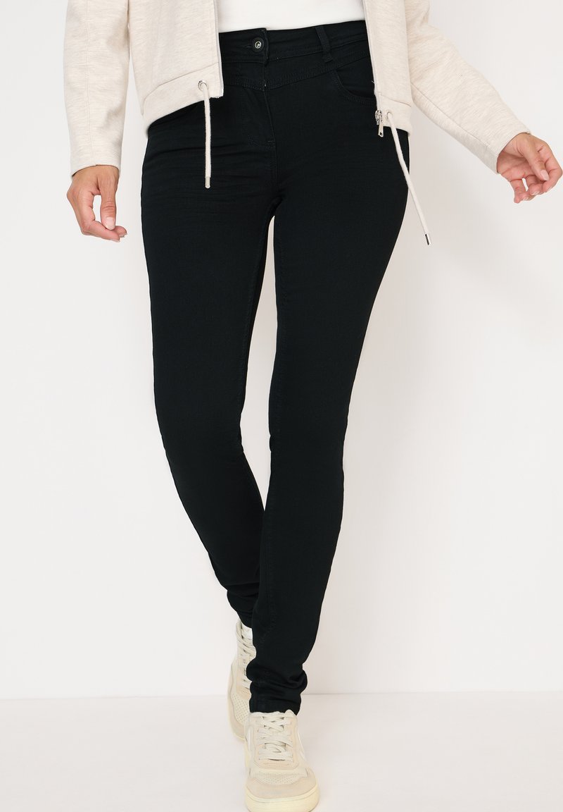 Jeans ajustados negros de denim elástico, con cintura alta, bolsillos frontales y una textura suave, combinados con zapatillas de color claro.