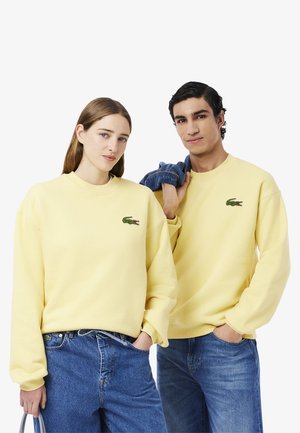Yellow cotton sweatshirts feature a crew neckline and the Lacoste crocodile logo. Paired with blue denim jeans.