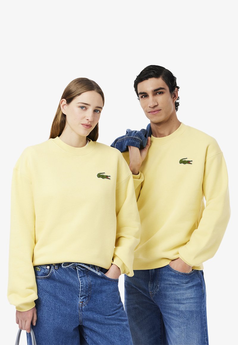 Gele katoenen sweatshirts hebben een ronde halslijn en het Lacoste krokodillenlogo. Combinatie met blauwe denim jeans.
