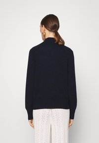 Granatowy sweter wełniany z wysokim kołnierzem i ściągaczem, z długimi rękawami i miękką fakturą. Tło jest czysto białe.