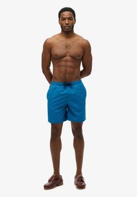 Blauwe zwembroek van lichtgewicht materiaal, met een elastische tailleband en een koord, en een subtiele logo-detail aan de zijkant.