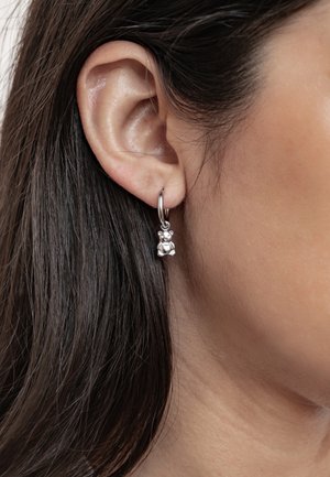 Ohr mit silbernem Creolen-Ohrring und kleinem Teddybär-Anhänger, mit dunklem Haar und heller Haut sichtbar.