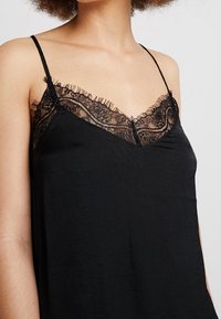 Camisole noire à fines bretelles et bordure en dentelle le long du décolleté. Texture lisse, design féminin avec des détails transparents au niveau de la poitrine.