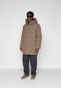 Lang brun parka med hætte, flere frontlommer og lynlås detaljer, parret med mørke cargo-bukser og sportssko.