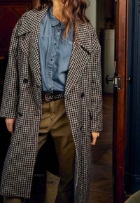 Trench coat a motivo pied-de-poule in bianco e nero, con un design doppiopetto e ampi revers, abbinato a una camicia di jeans e pantaloni khaki.