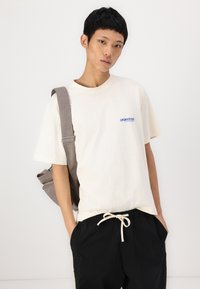 Beige bomulds t-shirt med korte ærmer, med et blåt logo. Båret med sorte drawstring bukser og en grå skuldertaske.