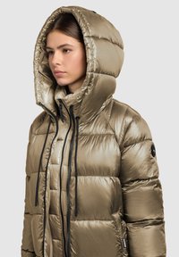 Gewatteerde jas met capuchon in metallic olijfgroene kleur, met een hoge kraag, zwarte ritsen en trekkoorden, en een doorgestikte textuur over de hele jas.