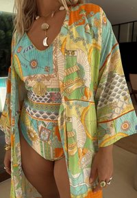 Sieviete valkā krāsainu rakstainu halātu un atbilstošu peldkostīmu ar gliemežvāku un pusmēness kaklarotu, gredzeniem un aprocēm telpās pie baseina.