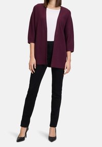 Žena v bílém topu, otevřeném fialovém kardiganu s 3/4 rukávy, černých slim kalhotách a černých lodičkách s špičatou špičkou, stojící na bílém pozadí.