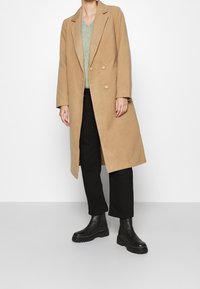 Beige kaksirivinen takki, jossa on viistot kaulukset ja napit; päällä vihreä neulepaita ja mustat lyhytlahkeiset housut, yhdistettynä mustiin saappaisiin.