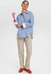 Blue and white striped button-up shirt with rolled cuffs, paired with beige linen trousers and red sneakers, standing against a neutral background.