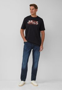 Schwarzes Grafik-T-Shirt mit "JAWS"-Design, kombiniert mit blauen Jeans und weißen Sneakers. Lässiger Schnitt, kurze Ärmel und weiche Stoffqualität.