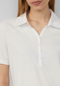 Weißes Poloshirt mit kurzen Ärmeln und Kragen, ausgestattet mit einer Knopfleiste und glatter Textur. Dezenter Glanz mit klassischen Designelementen.