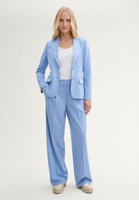 Světlomodrý přizpůsobený blazer a kalhoty širokého střihu vyrobené z hladkého materiálu; obsahuje dvě přední kapsy a bílý top pod ním.