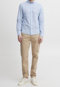 Vaaleansininen napitettava paita kevyesti teksturoitua kangasta, yhdistettynä beigeihin chinohousuihin ja valkoisiin tennareihin. Paidassa on pieni logo.