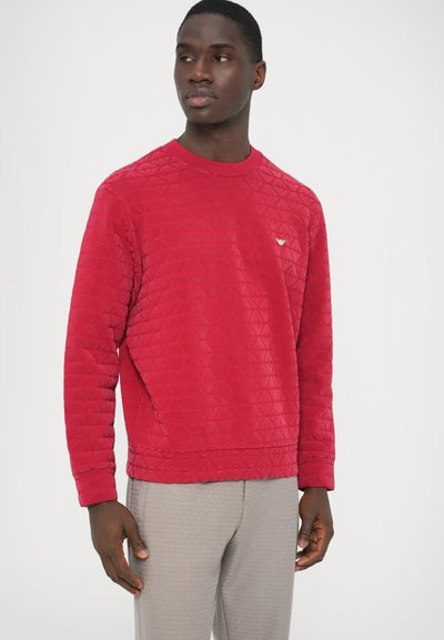 Κόκκινο καπιτονέ φούτερ με μακριά μανίκια, στρογγυλή λαιμόκοψη, διακριτική λεπτομέρεια λογότυπου στο στήθος και υφή φινιρίσματος. Συνδυασμένο με ανοιχτό γκρι παντελόνι.