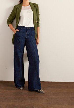 Vrouw draagt olijfgroene blazer, wit topje met zakken, wijde donkerblauwe jeans en puntige schoenen van slangenleer, staat met handen in de zakken.