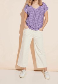 Top viola a maniche corte con scollo a V e motivo geometrico, abbinato a pantaloni a gamba larga color crema e sneakers bianche.