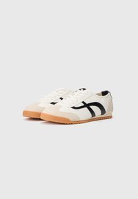White and beige sneakers with black stripes, textured fabric, and a rubber sole. Features round toe and lace-up front.
