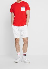 Červené bavlněné tričko s krátkým rukávem a bílou náprsní kapsou, doplněné bílými šortkami, bílými teniskami a pruhovanými ponožkami.