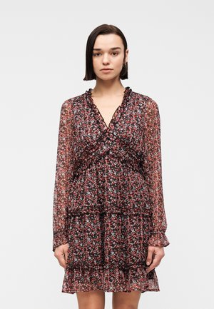 Jonge vrouw met steil donker haar, gekleed in een zwarte bloemenjurk met lange mouwen en ruches, tegen een effen lichte achtergrond.