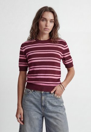 Jeune femme aux cheveux ondulés portant un pull à manches courtes rayé bordeaux et rose et un jean taille haute bleu clair, debout devant un fond blanc.