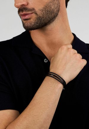 Uomo con indosso una maglietta nera a maniche corte e un braccialetto intrecciato in pelle nera, con la mano destra appoggiata sul petto, su sfondo grigio.