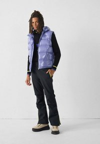 Lichtpaarse gewatteerde bodywarmer met capuchon, gecombineerd met een zwart shirt en zwarte broek met gele accenten, en beige wandelschoenen.