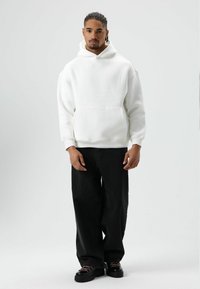 Witte hoodie met capuchon en een buidelzak, gecombineerd met zwarte wijde jeans en zwarte schoenen met roze veters, geplaatst tegen een grijze achtergrond.