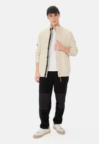 Cremefarvet tekstureret cardigan med sorte knapper, båret over en hvid skjorte. Sorte bukser med farveblokkerede paneler, parret med hvide sneakers.