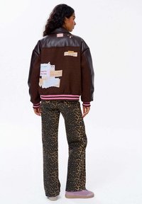 Kobieta z kręconymi ciemnymi włosami nosi brązową kurtkę z łatami, spodnie w panterkę i różowe trampki, stojąc na tle białym.
