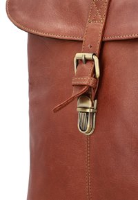 Smeđi kožni ranac s zakrivljenim poklopcem, mesinganim hardverom i detaljnim šavovima. Upadljivi kopča s zaobljenim dizajnom i podesivim remenom.