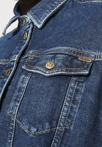 Jeanshemd in Mittelblau mit klassischem Kragen, Brusttaschen, goldfarbener Hardware und kontrastierendem Nähen entlang der Nähte.