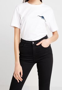 Valkoinen puuvillainen t-paita, jossa on tummansininen dinosauruksen siluetti vasemmassa rintakohdassa. Yhdistetty korkeavyötäröisiin mustiin skinny-farkkuihin.