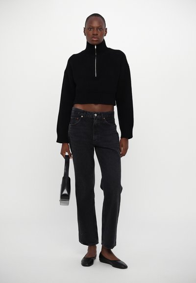 Black cropped sweater with high collar and silver zipper, paired with dark straight-leg jeans and flat black shoes, holding a black bag.