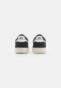 Pari musta ja valkoisia New Balance -tenniskenkiä näkyy takaa valkoista taustaa vasten, korostaen brändin logoa kantapäässä.
