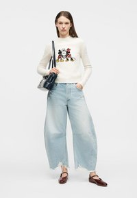 Femme portant un pull blanc avec un imprimé Mickey et Minnie, un jean large bleu clair déchiré, des chaussures marron, et tenant un sac à main noir.