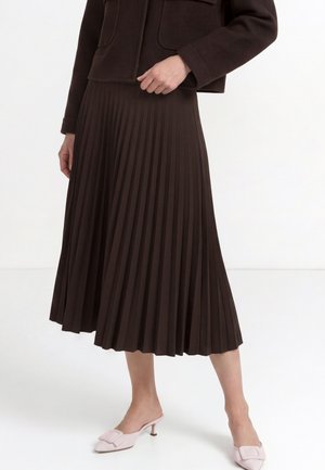 Mujer con una falda midi plisada de color marrón oscuro, chaqueta oscura y mules rosa claro con tacón bajo sobre fondo blanco.