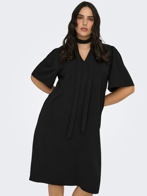 Femme aux longs cheveux foncés portant une robe noire longueur genoux avec de larges manches courtes et un foulard assorti noué autour du cou, debout les mains sur les hanches.