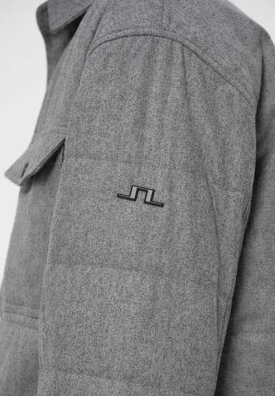 Harmaa villatakki, jonka hihais on minimalistinen musta geometrinen logo ylävartalon lähellä ja sivussa läpällinen tasku.