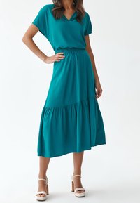 Robe teal à manches courtes avec un décolleté en V, taille élastique et jupe à volants. Tissu doux avec une silhouette fluide, assortie à des sandales à talons blocs blanches.