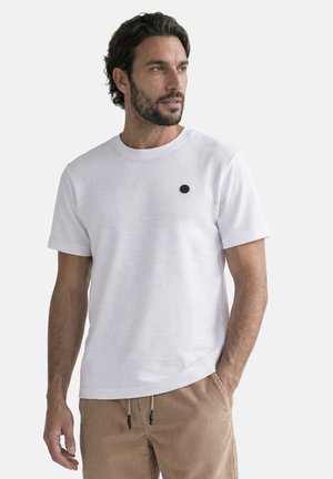 Homme aux cheveux foncés et à la barbe portant un t-shirt blanc uni à manches courtes et un pantalon en velours côtelé marron clair, regardant légèrement sur le côté.