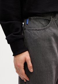 Grey denim jeans with a subtle texture, featuring a blue "HUGO" tag and standard pockets, paired with a black sweatshirt at the cuff.