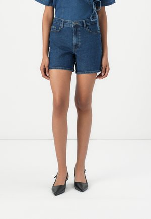 Vrouw die een donkerblauwe denim shorts met hoge taille en zwarte slingback-hakken met puntige neus draagt, staand tegen een effen witte achtergrond.