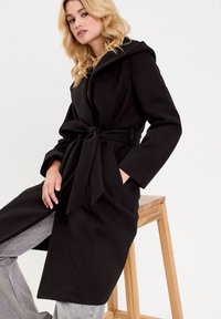 Manteau noir en mélange de laine avec une taille ceinturée, manches longues et capuche. Texture lisse et conception jusqu'aux genoux. Modèle assis sur un tabouret en bois.