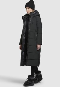 Frau, die einen langen, schwarzen, gepolsterten Mantel mit Kapuze, eine schwarze Mütze, Leggings und schwarze isolierte Winterstiefel trägt und vor einem hellgrauen Hintergrund steht.